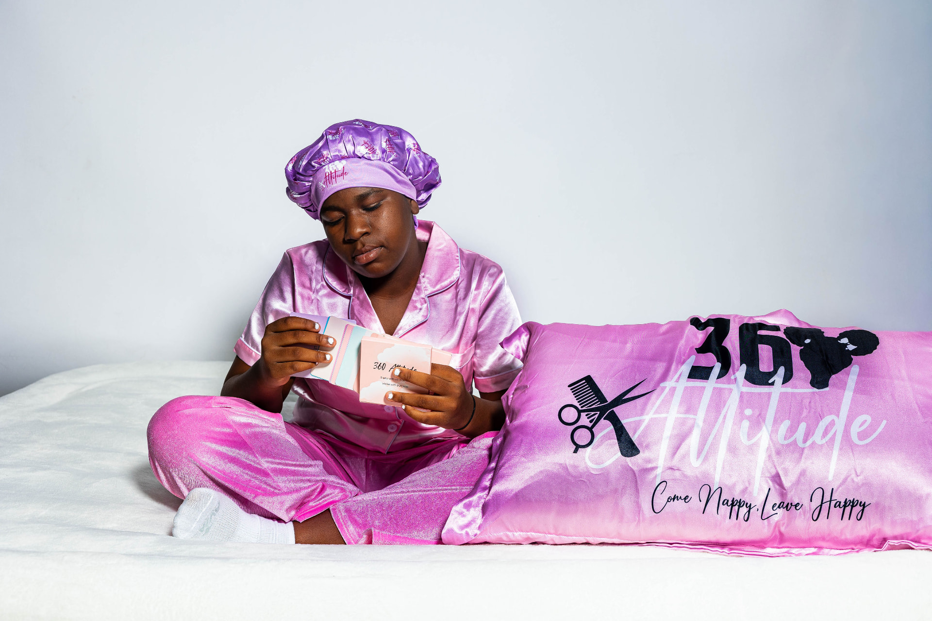 Girl in pink satin pajamas and bonnet reading 360 Attitude affirmation cards beside a pink logo pillowcase
